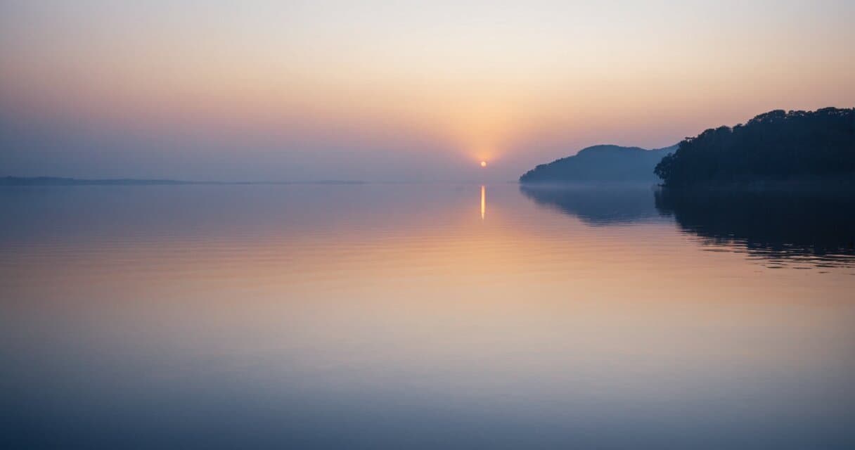 A calm night sky over still water with a single soft glow on the horizon